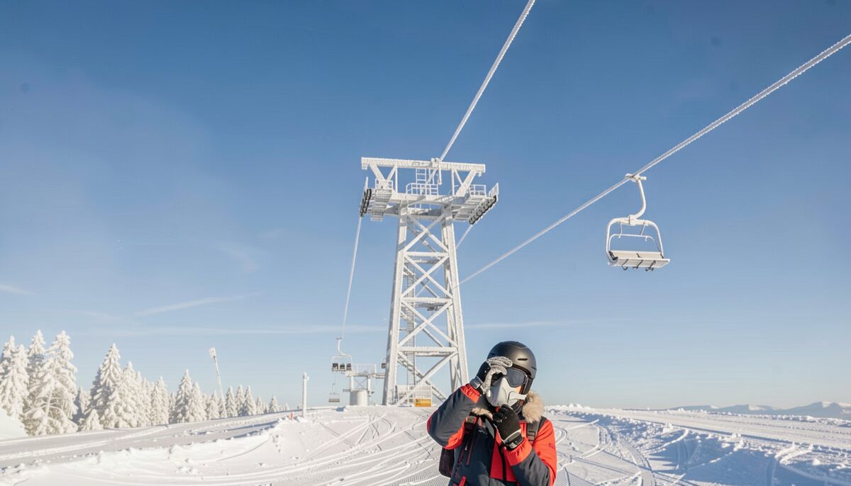 A frost-covered lift tower on a clear blue morning with a skier below adjusting a face mask.